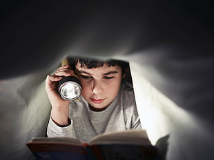 child under the white bed sheet holding flashlight while reading book HD wallpaper